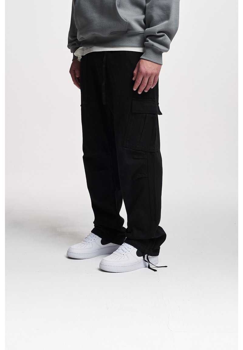 Personne portant un pantalon cargo noir, un sweat-shirt gris et des baskets blanches, debout sur un sol blanc devant un fond uni.