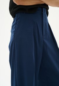 Marineblaue Hose mit glatter Textur, ausgestattet mit Falten und Seitentaschen, im entspannten Schnitt und mit mittelhoher Taille gestaltet.