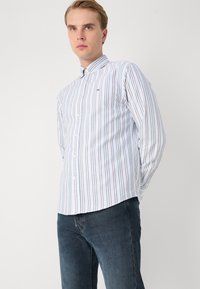 Hemd mit Button-Up-Auschnitt und vertikalen Streifen in Blau, Weiß und Pastellfarben; verfügt über einen Kragen und ein Logodetail auf der Brust. Kombiniert mit dunklen Jeans.