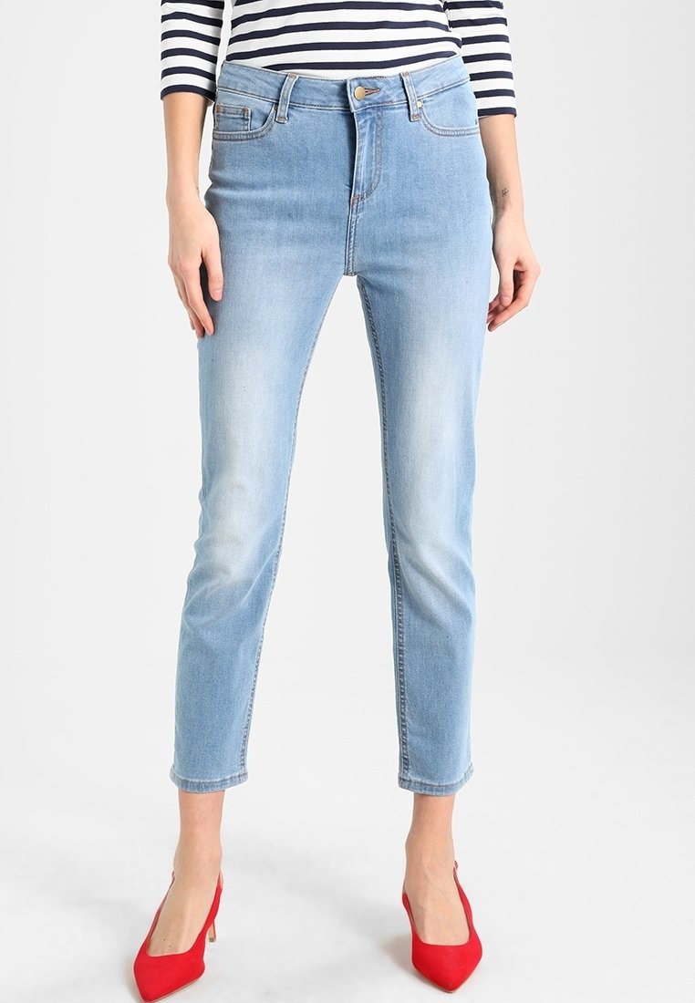 Jeans de tiro alto en azul claro con ajuste slim y ligero desvanecido. Presenta cinco bolsillos y un lazo para cinturón, combinados con tacones altos rojos.