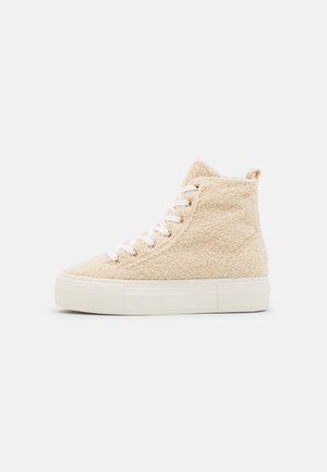 Baskets montantes beige avec dessus en polaire texturée, lacets blancs et épaisse semelle en caoutchouc blanc sur fond uni.
