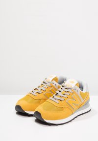 Ett par gula och grå New Balance 574-sneakers med vita sulor, grå snören och svart mönstring mot en vit bakgrund.