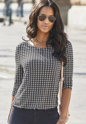 Schwarz-weiß gemusterte Bluse mit Dreiviertelärmeln, rundem Ausschnitt und einem kleinen Schlüssellochdetail; kombiniert mit marineblauen Jeans und Sonnenbrille.