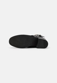 Chaussure habillée en cuir noir avec un petit talon carré, une bride à boucle, et une semelle en caoutchouc texturée, vue du dessous.