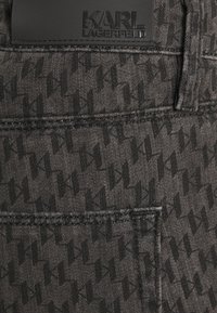Pantalones de mezclilla gris con un sutil patrón geométrico negro y una etiqueta de marca negra rectangular en la cinturilla.