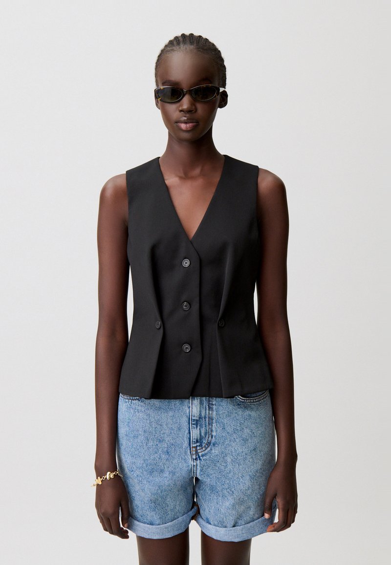 Top nero senza maniche con scollo a V e chiusura doppiopetto con bottoni, abbinato a pantaloncini di denim con orlo arrotolato. La texture appare liscia e su misura.