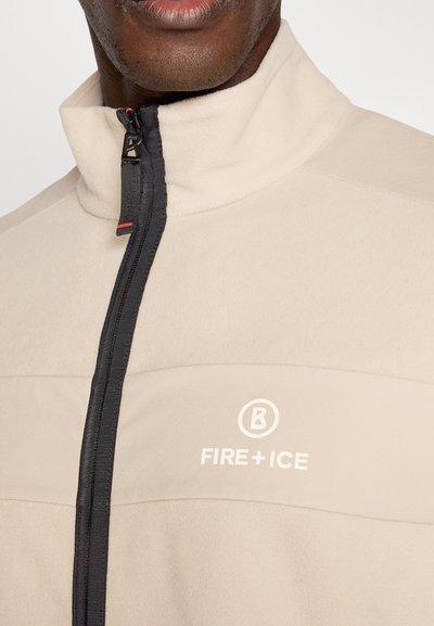 Nahaufnahme einer Person, die eine beige Reißverschlussjacke mit schwarzem Reißverschluss und weißem "FIRE + ICE"-Logo auf der Brust trägt.