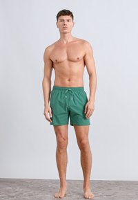 Zelené plavkové šortky s pásom na sťahovanie, z ľahkej tkaniny a s hladkou textúrou, prezentované postavou stojacou na jednoduchom pozadí.