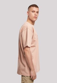 Světle růžové bavlněné tričko s krátkými rukávy, kulatým výstřihem a tištěnými grafikami na zadní straně. Tkanina vypadá měkce a má hladkou texturu.