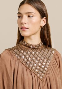 Vestido beige de cuello alto con una tela texturizada. Presenta intrincados bordados de cuentas y lentejuelas en un patrón de diamante en el cuello.