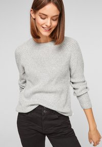 Pull en maille gris clair avec un col ras du cou, texture côtelée et manches trois-quarts retroussées, associé à un pantalon noir ajusté.