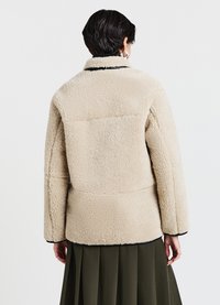 Veste polaire beige avec une texture douce, un col avec col, et des accents noirs, présentant une coupe décontractée et des coutures aux épaules.