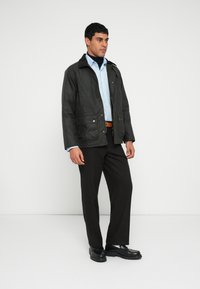 Tmavě zelená nepromokavá bunda s límcem, zapínáním na patentky a předními kapsami, nošená přes světle modrou košili a tmavé kalhoty. Černé boty.