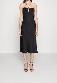 Černé saténové midi šaty s upnutým korzetem, detailem ve tvaru klíče a tenkými ramínky. Hladká textura a splývavá sukně. Kombinováno s černými sandály na vysokém podpatku.