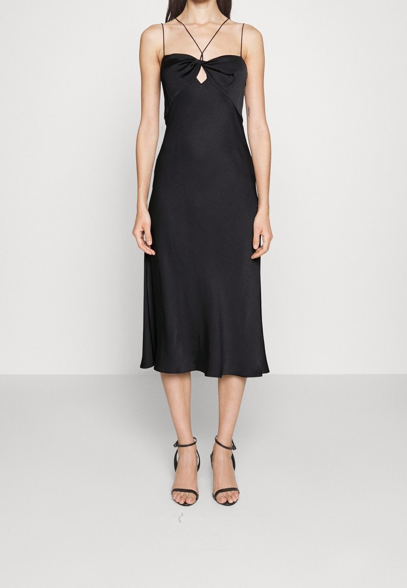 Černé saténové midi šaty s upnutým korzetem, detailem ve tvaru klíče a tenkými ramínky. Hladká textura a splývavá sukně. Kombinováno s černými sandály na vysokém podpatku.