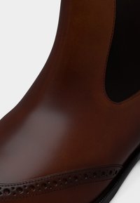 Chaussure en cuir marron lisse avec des détails en brogue près de la semelle et un panneau latéral élastique, mettant en valeur un savoir-faire et un design de qualité.