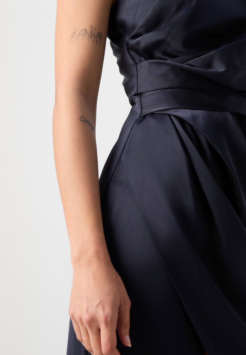 Granatowa satynowa sukienka z plisowanym wzorem, z grubym paskiem w talii. Gładka tekstura i subtelny połysk, z widocznymi tatuażami na ramieniu.