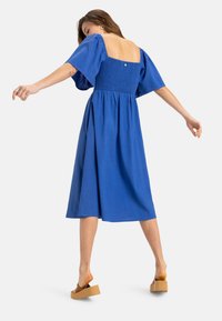 Vestido midi azul con corsé fruncido, mangas cortas abullonadas y espalda abierta. Combinado con sandalias de plataforma marrones.