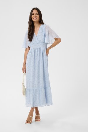 Femme souriante portant une robe maxi bleu clair à pois avec des manches volantées, tenant un sac à main beige et portant des sandales blanches.