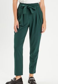 Pantalons verts taille haute avec une ceinture nouée, design plissé, jambes fuselées et texture lisse. Associés à des mocassins noirs épais.