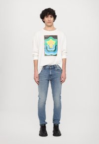 Hvit langermet skjorte med et fargerikt geometrisk grafisk design; sammen med lyseblå jeans og svarte sko.