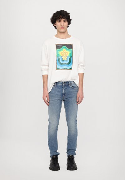 Witte langemouwen shirt met een kleurrijk geometrisch grafisch ontwerp; gecombineerd met lichtblauwe jeans en zwarte schoenen.