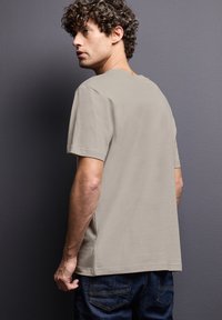 Ljusbeige t-shirt med kort ärm och avslappnad passform. Materialet verkar mjukt med en slät yta. Bärs med mörka jeans; synlig design på ryggen.