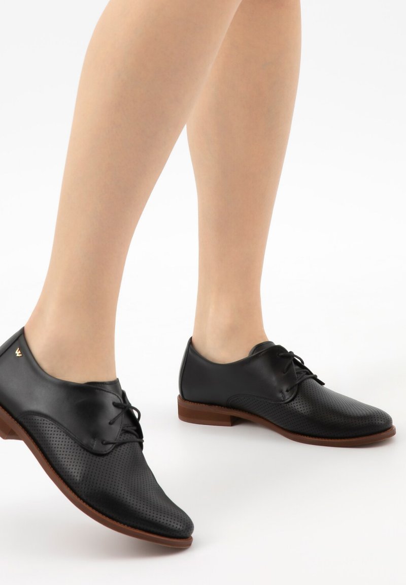 Czarne, perforowane, skórzane buty sznurowane z brązowymi podeszwami, noszone na bose nogi na białym tle.