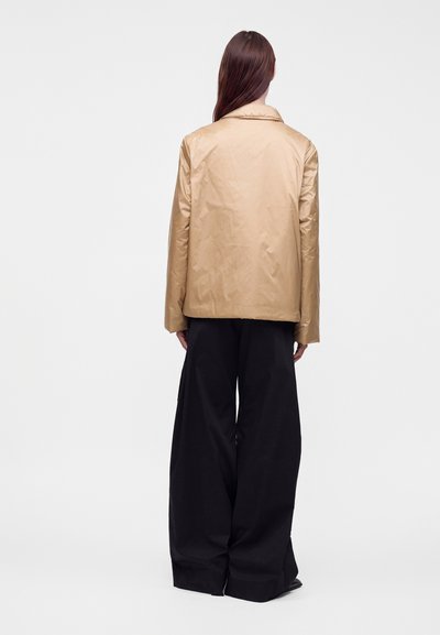 Femme aux longs cheveux bruns portant une veste beige et un pantalon noir à jambes larges, de dos, sur un fond clair uni.