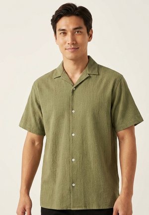 Homme portant une chemise texturée vert olive à manches courtes et boutonnée, debout devant un fond clair uni.