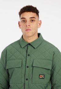 Gequiltede groene jas met knoopsluiting, twee borstzakken, contrasterende logopatch en een klassiek kraagontwerp.