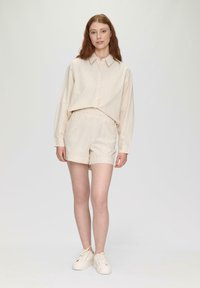 Cremfarbendes Hemd mit Knopfleiste, das einen Kragen und lange Ärmel hat, kombiniert mit passender Shorts, beides aus einem leichten, strukturierten Stoff. Weiße Sneakers.