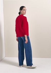 Roter Strickpullover mit langen Ärmeln, kombiniert mit hoch taillierten blauen Jeans im weiten Schnitt und weißen Sneakers, vor einem neutralen Hintergrund präsentiert.