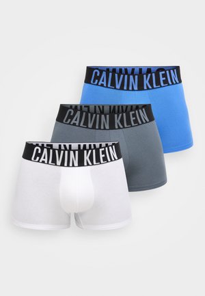 Três cuecas boxer masculinas Calvin Klein em branco, cinzento e azul, com cinturas pretas que exibem o nome da marca em letras grandes.
