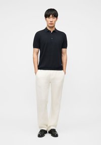 Junger Mann steht mit den Händen in den Taschen, trägt ein schwarzes Polohemd, weiße Hose und schwarze elegante Schuhe vor einem schlichten Hintergrund.