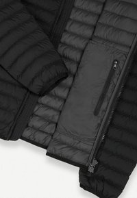 Chaqueta de plumas negra con textura acolchada, que presenta un interior suave, un bolsillo con cremallera y puños elásticos. El material parece ligero y aislante.