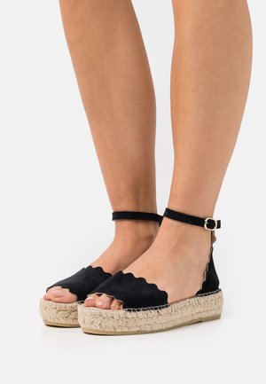 Feet wearing black scalloped-edge espadrille sandals with ankle straps and jute platform soles on a white background.