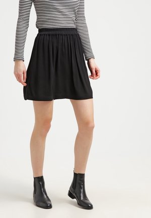 Femme portant une mini-jupe noire froncée, des bottines noires et un haut à manches longues rayé noir et blanc, debout sur un fond blanc.