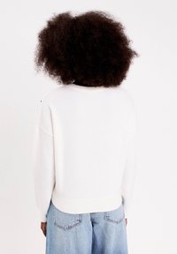 Personne avec des cheveux bouclés foncés portant un pull blanc et un jean en denim bleu clair, de dos sur un fond blanc uni.