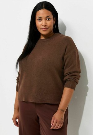 Mujer con cabello largo y negro que lleva un suéter marrón y pantalones a juego, de pie contra un fondo claro y liso, con pendientes de aro.