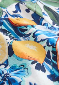 Tissu avec des motifs colorés de citrons et de fleurs bleues sur fond blanc, présentant des plis et des détails de texture.