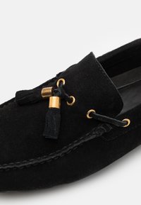 Mocassin en daim noir avec des détails cousus, doté de glands et d'accents métalliques dorés sur le lacet noué à l'avant.