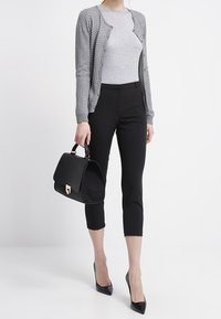 Grau gestrickte Strickjacke, eng anliegendes graues Oberteil, schwarze verkürzte Hose, schwarze Handtasche mit goldenen Beschlägen und schwarze spitze Pumps.