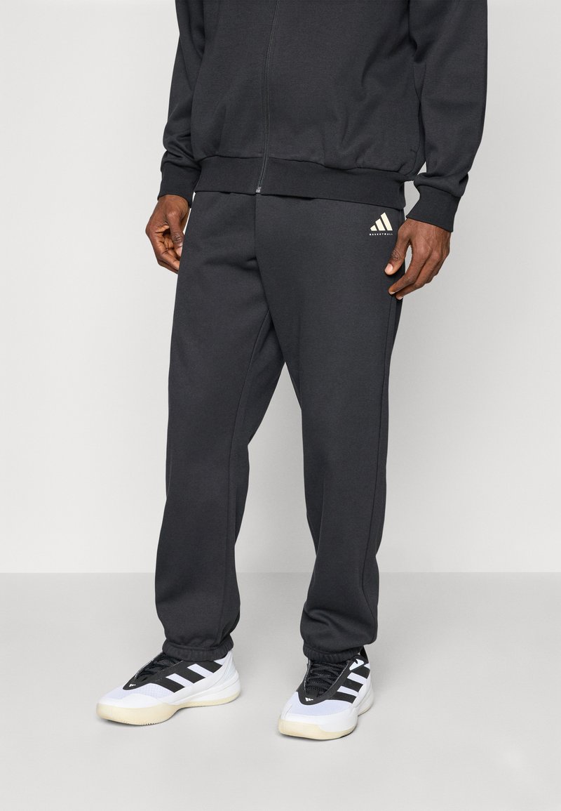 Schwarze Sporthose mit elastischem Bund, seitlichen Taschen und goldener Adidas-Logos auf dem linken Oberschenkel. Getragen mit weißen und schwarzen Sneakers.
