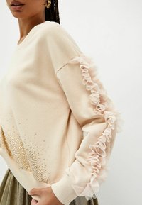 Mujer con un suéter beige con detalles de malla con volantes a lo largo de las mangas y cristales de oro cerca del dobladillo, combinado con una falda metálica plisada.
