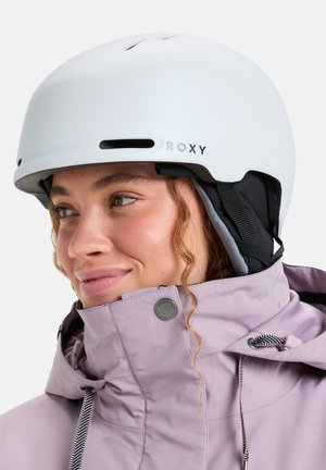 Jeune femme portant un casque blanc Roxy et une veste de ski lavande à col haut, regardant sur le côté avec un léger sourire.
