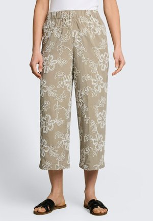 Frau trägt beige kurze Hosen mit weißer Blumenstickerei und schwarze Slip-on-Sandalen, steht vor einem einfarbigen Hintergrund.