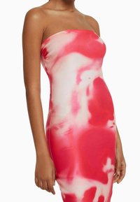 Femme portant une robe moulante sans bretelles avec un motif de tie-dye abstrait rouge et blanc, se tenant devant un fond blanc.