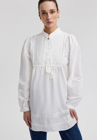 Blusa bianca con maniche a sbuffo, dotata di una chiusura con bottoni, frange e dettagli in pizzo intricati lungo il corpetto e le maniche.