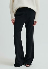 Kobieta w czarnych szerokich spodniach z wysokim stanem i białym swetrze z długim rękawem, stojąca boso na jednolitym tle.